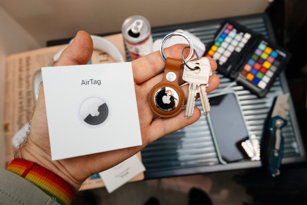Des clés reliées à un Apple AirTag, une boite d'Apple AirTag dans une main, sur un fond d'objets divers